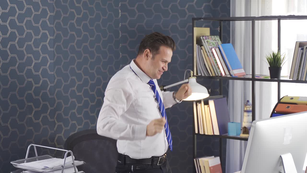 Cheerful businessman celebrating sitting at desk in office and working on computer with raised arms.