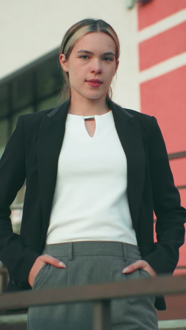 Confident lady in black blazer and white top poses with hands in pockets near iron railing in front of modern office building, exuding elegance, professionalism, and poise under clear sky