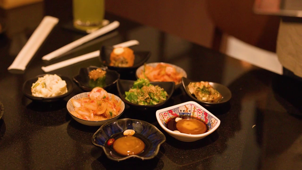 Uma pessoa organiza vários pratos pequenos de aperitivos japoneses em uma mesa escura de restaurante