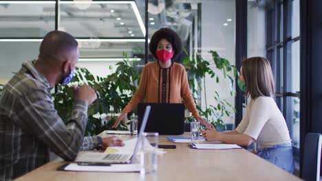 Diverse-business-people-wearing-face-masks-sitting-using-laptops-going-through-paperwork-in-office