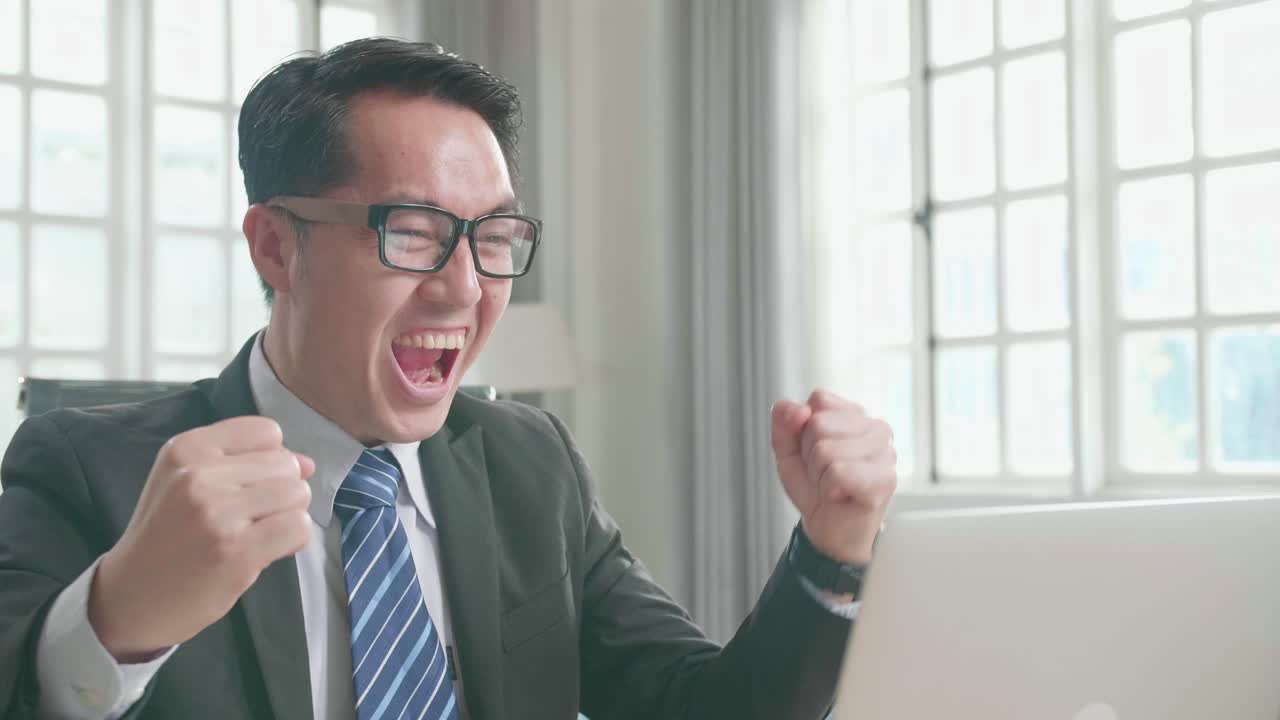 眼鏡をかぶったアジア人ビジネススーツを着てコンピューター画面を見て自宅で働いている間プロジェクトの入札で勝てて幸せです