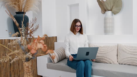 caucasian lady in white shirt and jeans takes laptop from coffee table. comfortable work conditions at home.
