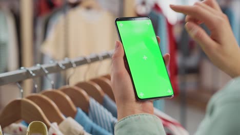 clothing store: female using smartphone with chroma key green screen display. clothes hanger with stylish branded items for retail sale in the background. close up footage of mobile device.