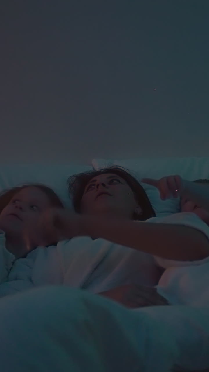 Mother and children point fingers at ceiling with demonstration stars in different colors. Night light and entertainment before sleep slow motion