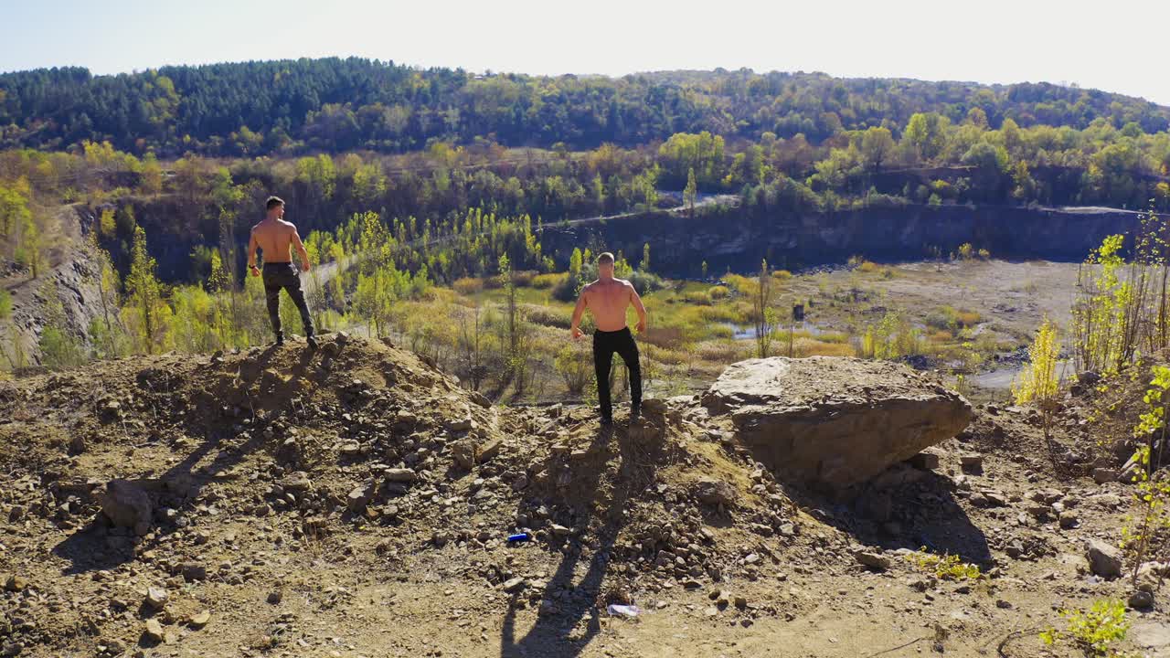 Backside view of two sportsmen on the rocky hill. Shirtless athletes showing their muscular body on the nature background in summer. Aerial view.