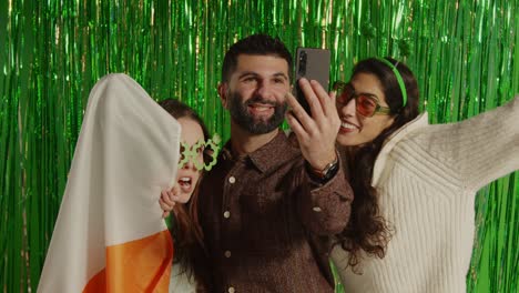 Studio-Shot-Of-Friends-Dressing-Up-With-Irish-Novelties-And-Props-Posing-For-Selfie-Celebrating-St-Patrick's-Day-Against-Green-Tinsel-Background-3