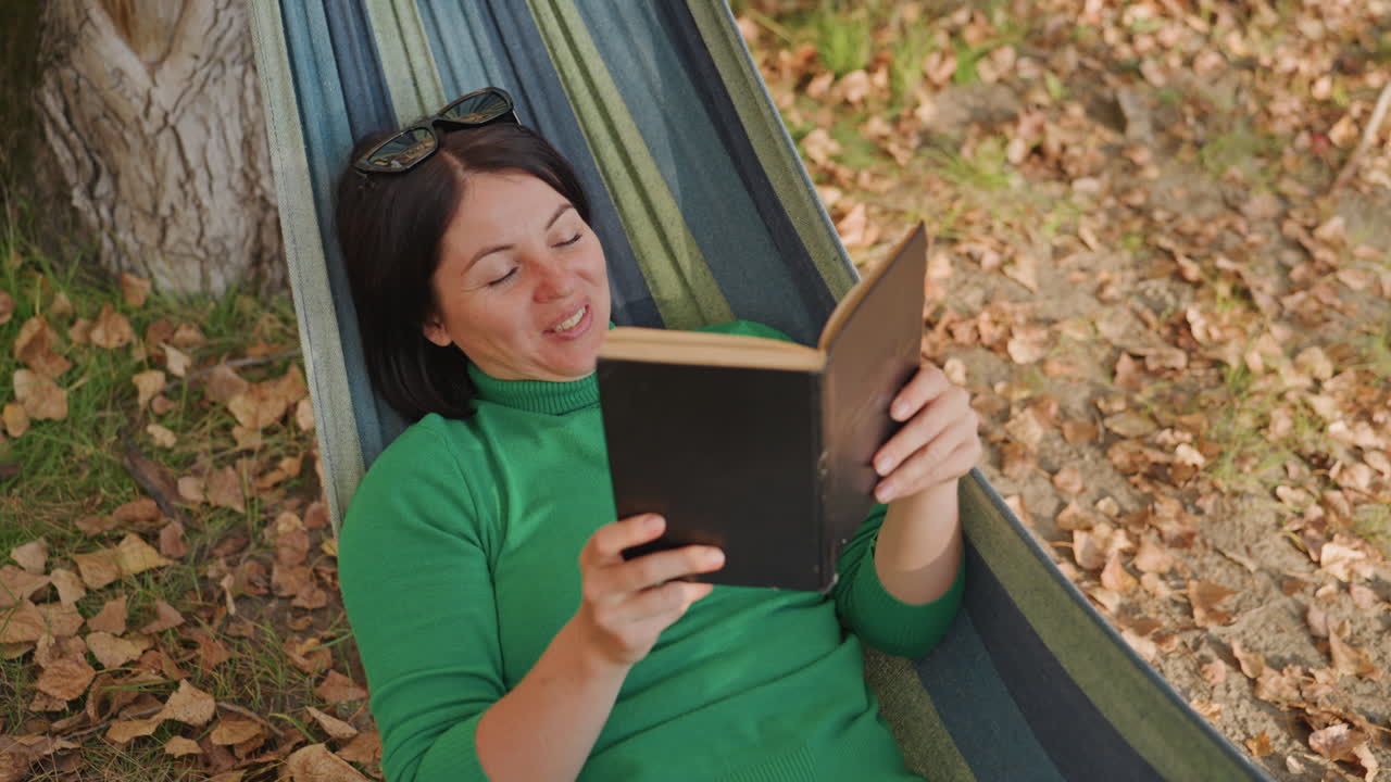 White woman hammock camping reads book, under tree among fallen leaves, green sweater, sunglasses on head, smiling and relaxed, student reader vibe, closeup on pages, warm golden light, peaceful