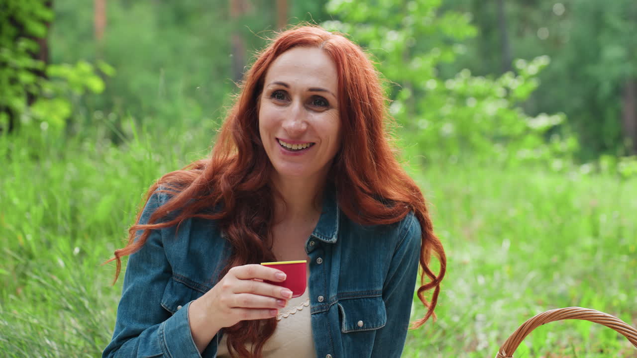긴 빨간 머리를 가진 가을을 사랑하는 사람이 따뜻한 차 컵을 들고, 녹색 숲으로 둘러싸인 야외에서 평화롭게 채팅하면서 따뜻한 피크닉 분위기 속에서 차를 마시기 전에 부드러운 바람을 불고 있어요.