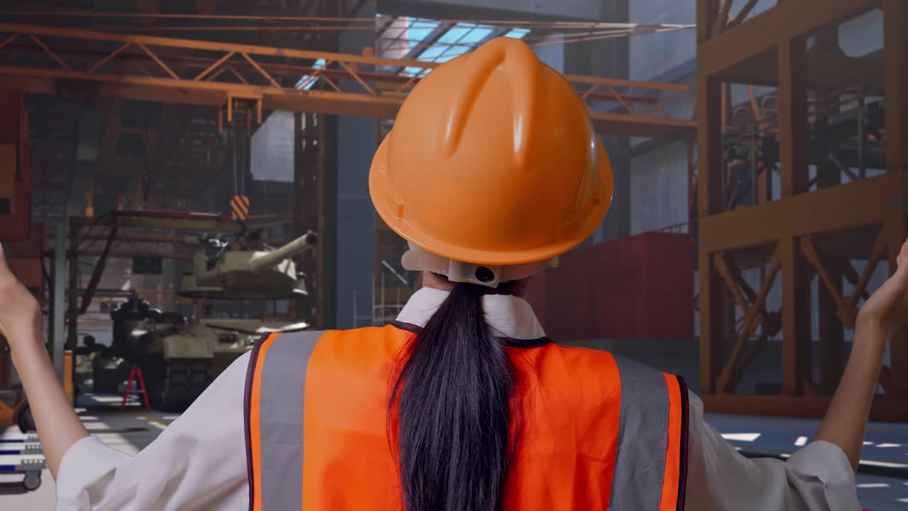 Primo piano da dietro di un'ingegnera con casco di sicurezza che allarga le braccia mentre è in piedi, carro armato militare in costruzione in uno stabilimento industriale