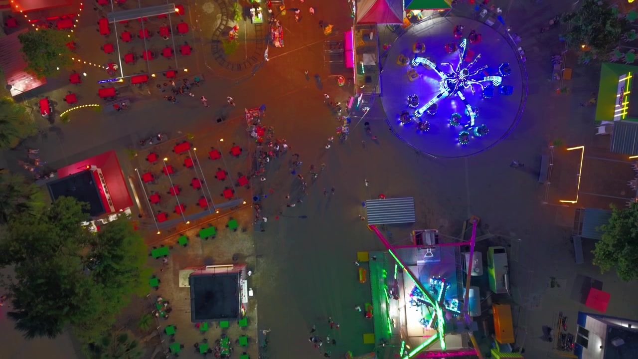 Top-down natlig luftdroneoptagelse, der flyver fremad over skarpt oplyste tivoliture, madområder og spilleboder i Santo Domingo Malecon forlystelsespark