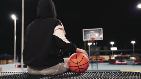 Vista-Trasera-De-Una-Jugadora-De-Baloncesto-Con-Capucha-Sentada-Y-Sosteniendo-La-Pelota-Mientras-Toma-Un-Descanso-Después-De-Su-Sesión-De-Entrenamiento-En-La-Cancha-Al-Aire-Libre-Por-La-Noche-1