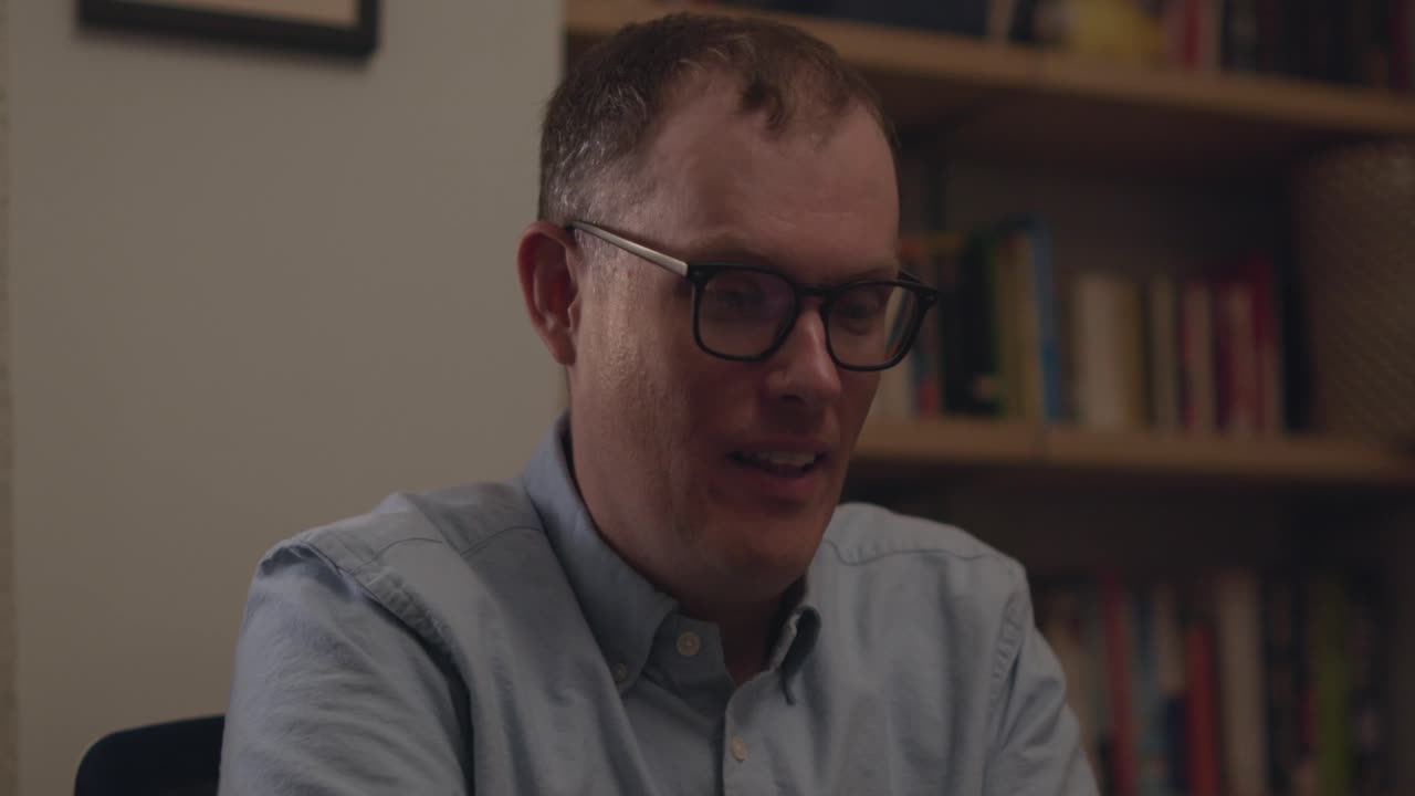 A man in glasses at work talks and laughs to someone off screen on a virtual call