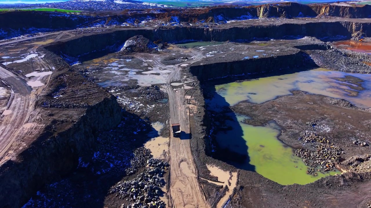 Haul truck in a deep quarry, Bulgaria. Wide aerial view the heavy vehicle moving on a central dirt path between green mineral pools and rocky terraces.