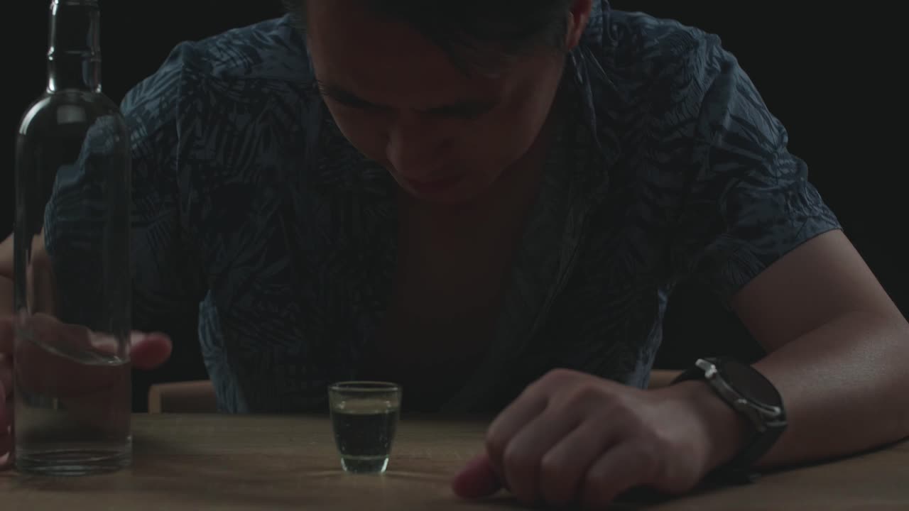 술에 취한 아시아인 남자가 술을 마시기 전에 글라스에 보드카를 부어 검은 바탕에서 잠을 자고 있습니다.