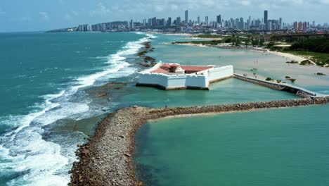 Dolly-in-aerial-drone-wide-shot-of-the-historic-star-shaped-Reis-Magos-fort-built-on-a-reef-with-the-coastal-capital-city-of-Natal-in-Rio-Grande-do-Norte,-Brazil-in-the-background-on-a-warm-summer-day