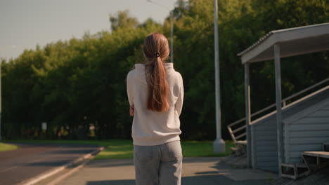 back view of young woman with long hair in casual hoodie, arms folded, walking outdoors by stadium seating on a sunny day, with electric poles and lush green trees in the background