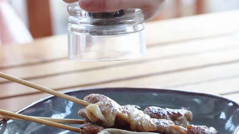close-up of seasoning being added to skewered meat on a dark plate at a wooden table.
