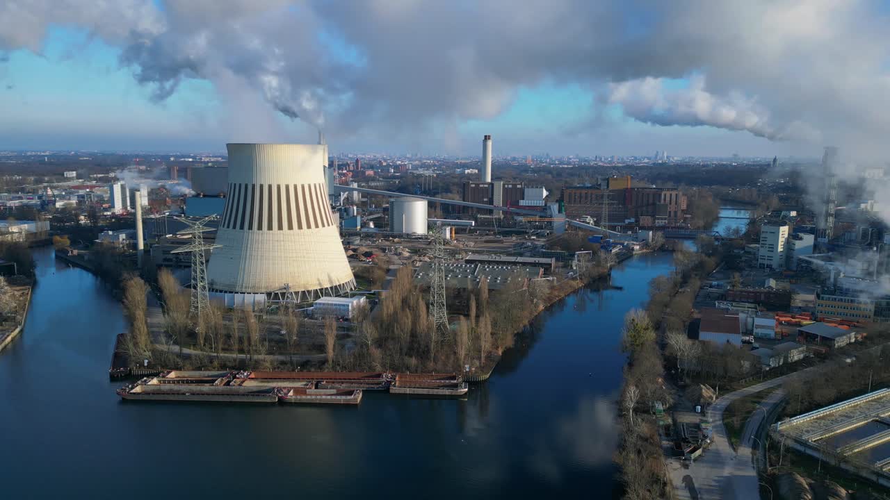 Aerial view of a thermal power plant emitting smoke on a sunny day, with a river and a city in the background. Magic aerial view flight static tripod hovering drone