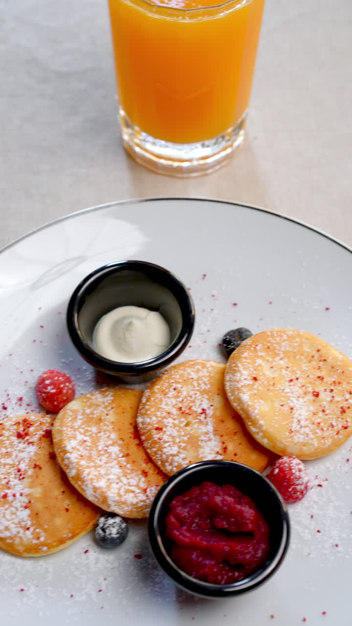 Traditional syrniki cheese pancakes lying on textured gray linen, garnished with ripe raspberries, dollop of sour cream, freshly squeezed orange juice