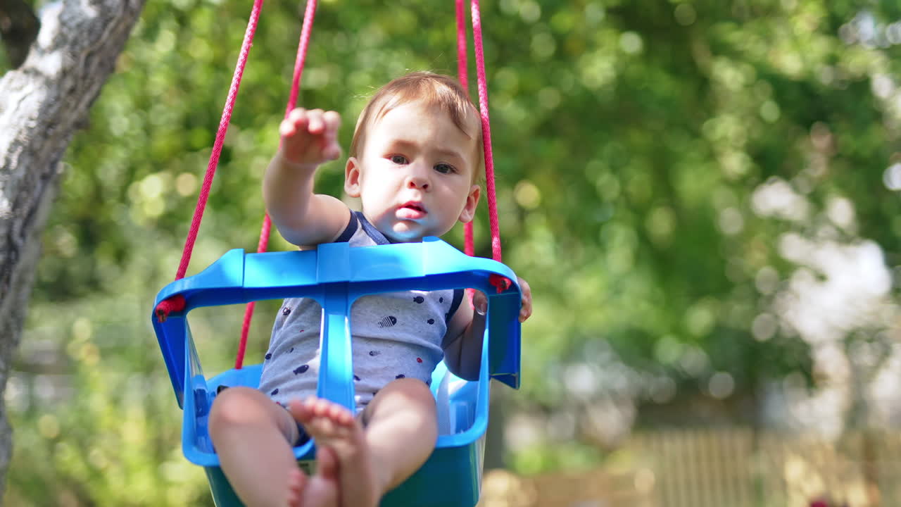 Adorable little kid sitting in a swing. Lovely calm boy waving his hand while swinging. Nature backdrop.