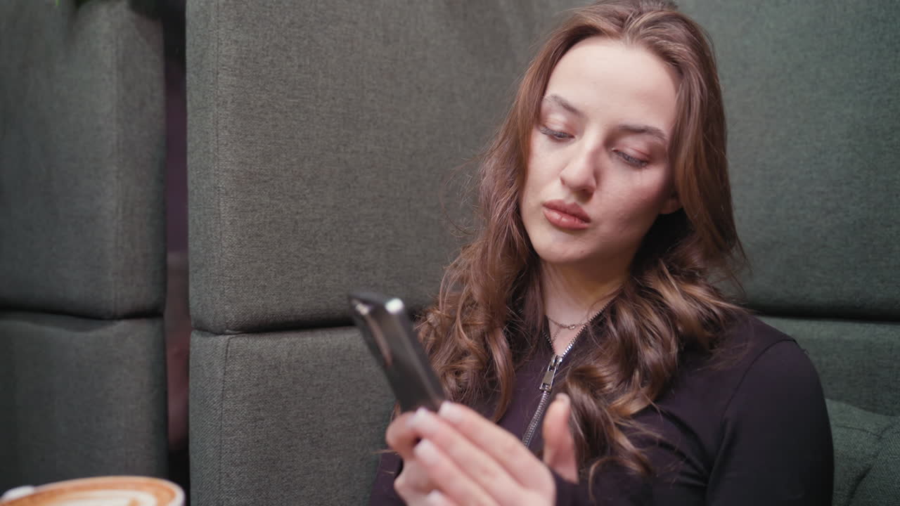 Young woman in black outfit seated in green booth fixated on smartphone with dessert and cup of latte on wooden table. Surrounded by lush indoor plants, and books