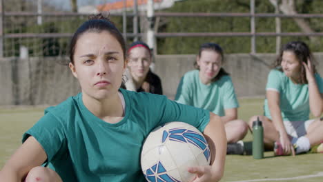 sporty teenaged girl holding football in front of camera, while her teammates relax behind