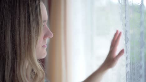 girl longingly places hand on inside of window while looking outside, close up