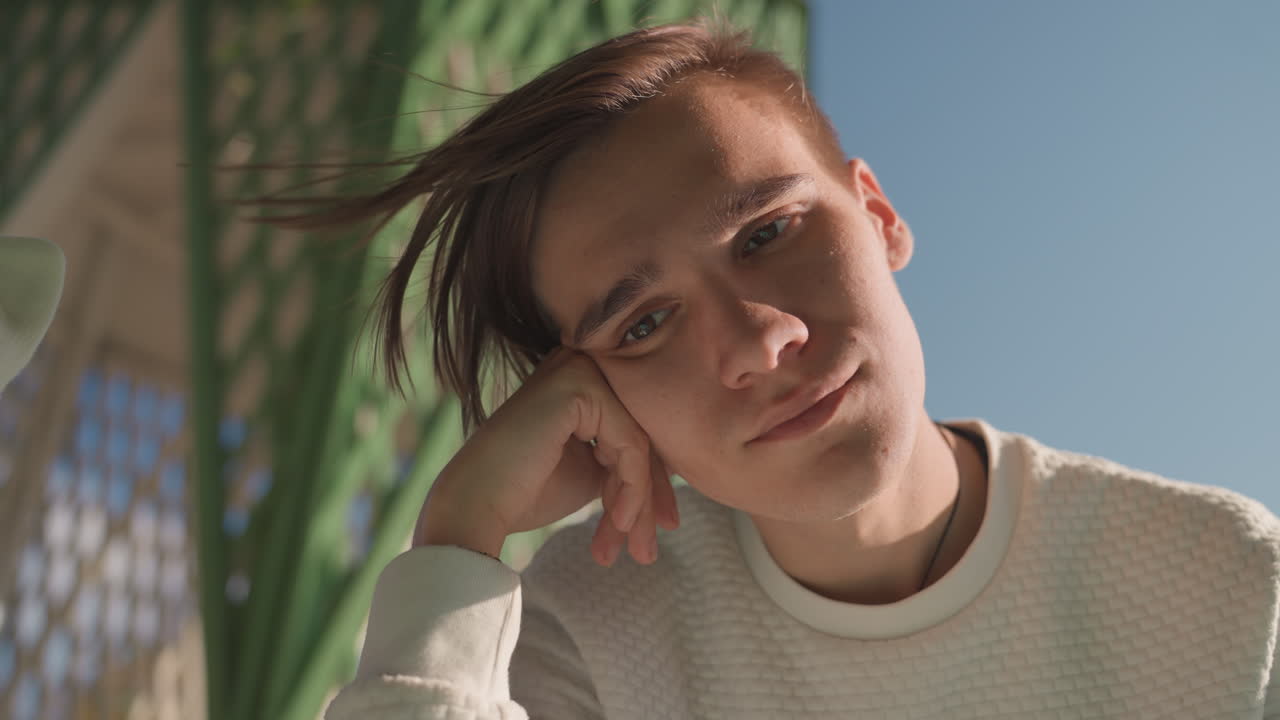 Resolute young white man outdoors, young teacher style resting head on hand, steady gaze toward camera with warm afternoon light, textured sweater and green architecture, quiet acceptance of future