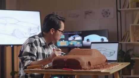 asian man automotive designer looking at a laptop and using rake or wire to smooth out the surface and create details in the sculpture of car clay in the studio