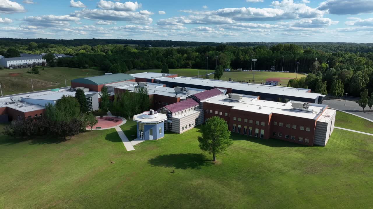 Aerial view of modern school building in Virginia, surrounded by open green fields and forests. Brick and glass architecture with a sports field in background. Closed high school in summer season