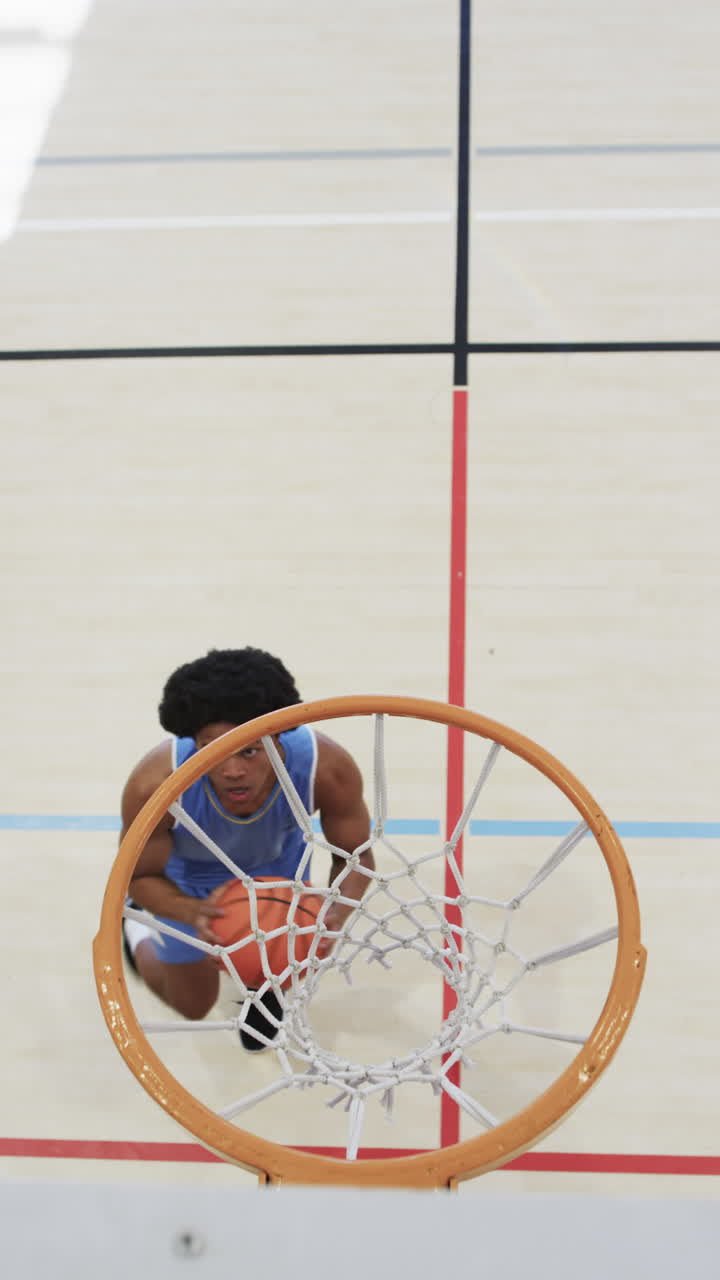 Vertical video of male african american basketball player training in indoor court, in slow motion