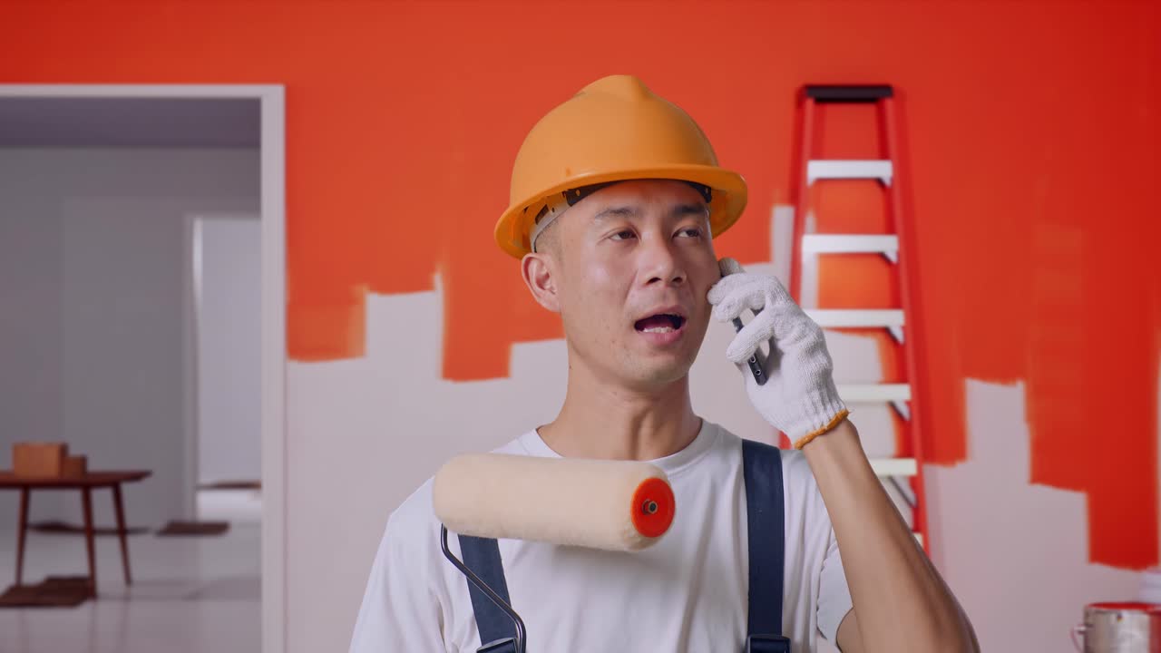 Close Up Of Asian Man Painter Wearing Safety Helmet Talking On Smartphone And Looking Around While Standing With Painting Wall In Room