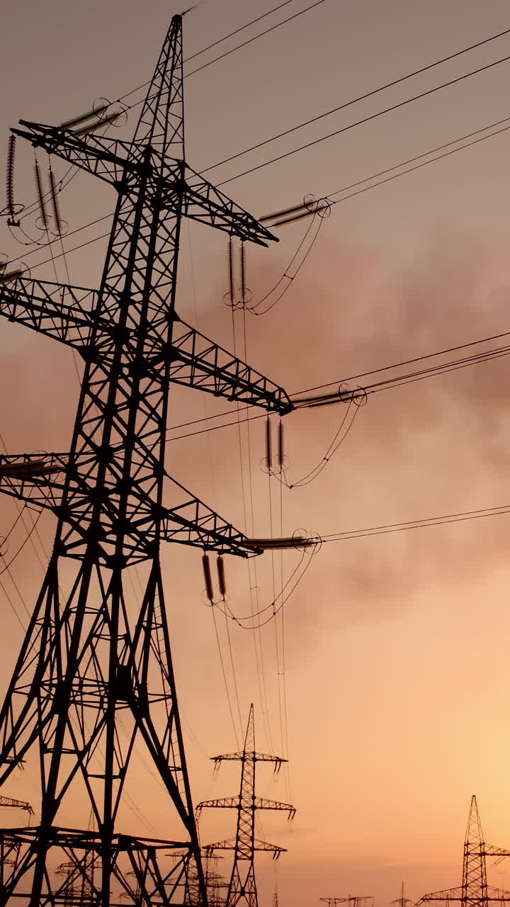 Electricity pylons against smoke stacks at sunset. High-voltage electric towers on the evening sky. Factory pipes with smoke pollute the air. Motion camera down. Vertical video