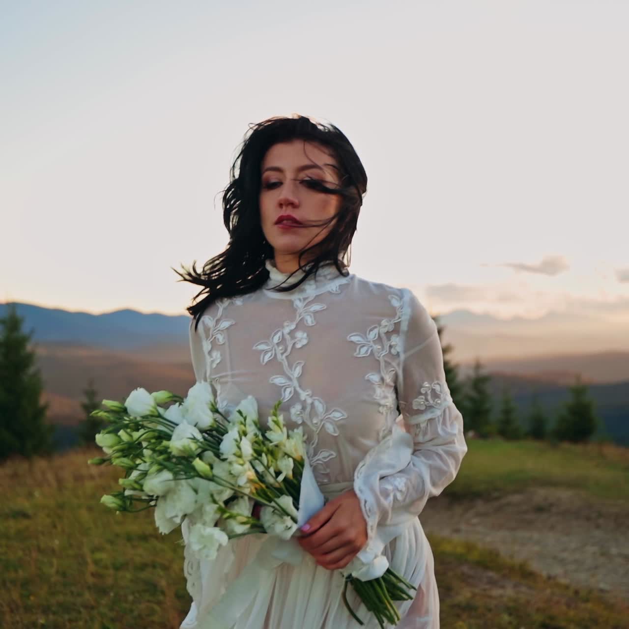 Una donna solitaria in abito bianco si avvicina alla telecamera, tenendo un bouquet tra le mani. Il vento le fa svolazzare i capelli e il vestito. Uno splendido paesaggio montano con il sole al tramonto fa da sfondo.