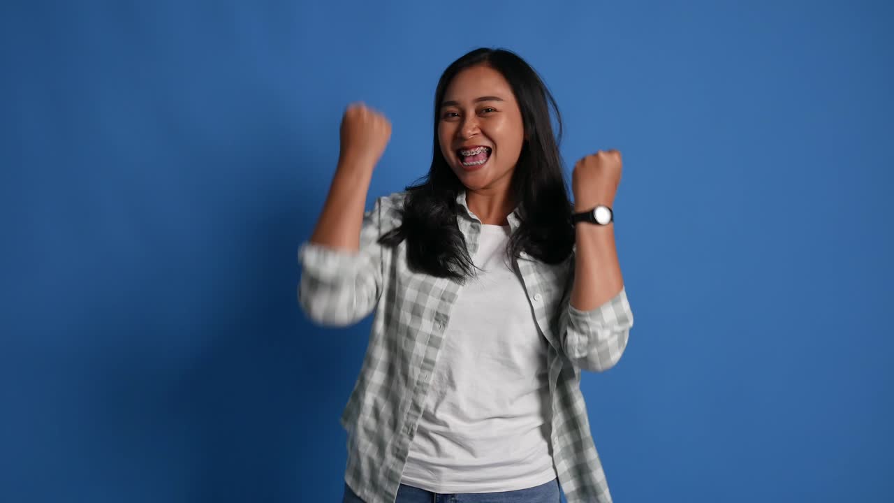Happy asian woman celebrating success with raised fists on blue background