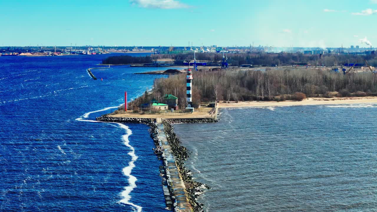 A tall black and white lighthouse stands at the end of a long stone jetty splitting vivid blue Baltic Sea from muted estuary waters, framed by forest and distant city skyline.
