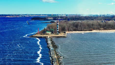 a tall black and white lighthouse stands at the end of a long stone jetty splitting vivid blue baltic sea from muted estuary waters, framed by forest and distant city skyline.