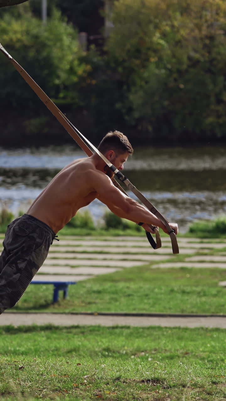 Young active man exercises outdoors with straps. Topless athlete doing workout on trx belts near the river in a summer day. Vertical video