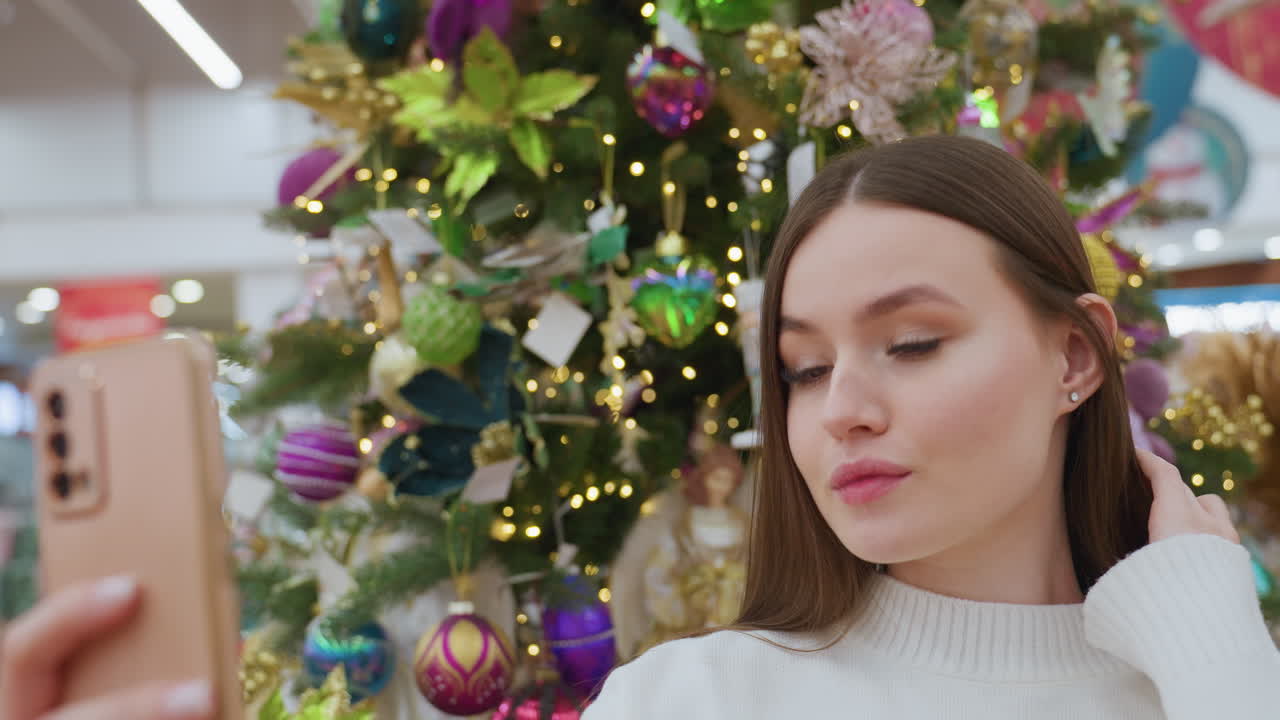 Beautiful lady in white sweater takes a selfie in front of vibrant Christmas tree in a well-lit decor store, she adjusts her hair while other shoppers are blurred in the background