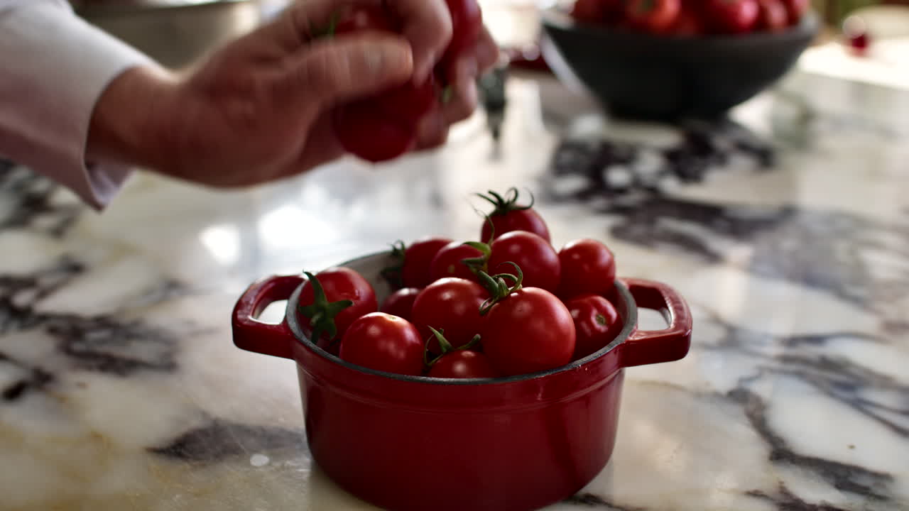 Close-up of a chef's hands taking fresh red cherry tomatoes from a small red pot in a generic kitchen. Professional culinary ingredient prep for a healthy dish