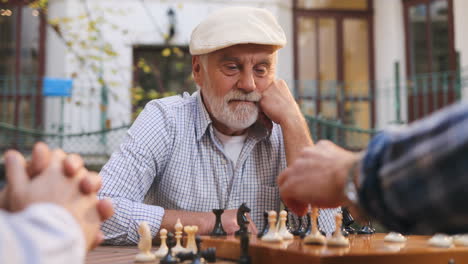 Portrait-Shot-Of-The-Grey-Haired-Good-Looking-Man-On-Retirement-Sitting-At-The-Table-On-A-Fresh-Air,-Leaning-On-His-Arm-And-Looking-At-The-Friend's-Hand-Moving-A-Chessman-On-A-Board