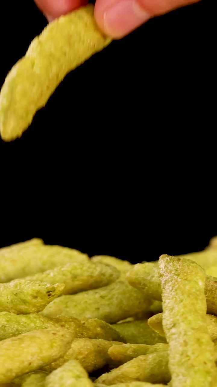 Uma mão pega em uma pilha de salgadinhos de ervilha verde contra um fundo preto.