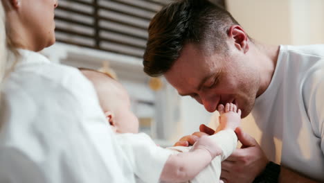 mid-aged caucasian man is kissing happily the feet and toes of his newborn son. newly-made mom and dad rejoicing their first baby. close up.