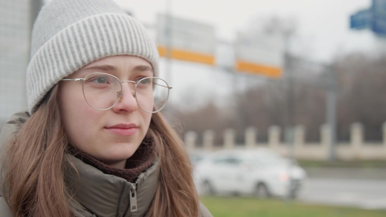 Primo piano di donna bianca con occhiali e berretto di maglia, studentessa urbana che aspetta a bordo strada con bokeh morbido ed espressione riflessiva, dettaglio chiaro degli occhiali, tonalità della pelle naturali e leggero movimento.
