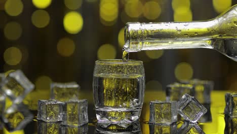 barman pour frozen vodka from bottle into shot glass. ice cubes against shiny gold party background