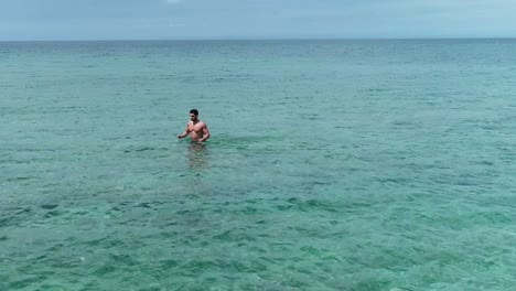 young handsome caucasian strong man wearing bathing suit walking in blue ocean water on cloudy day, circle aerial, palm island, tripoli, lebanon