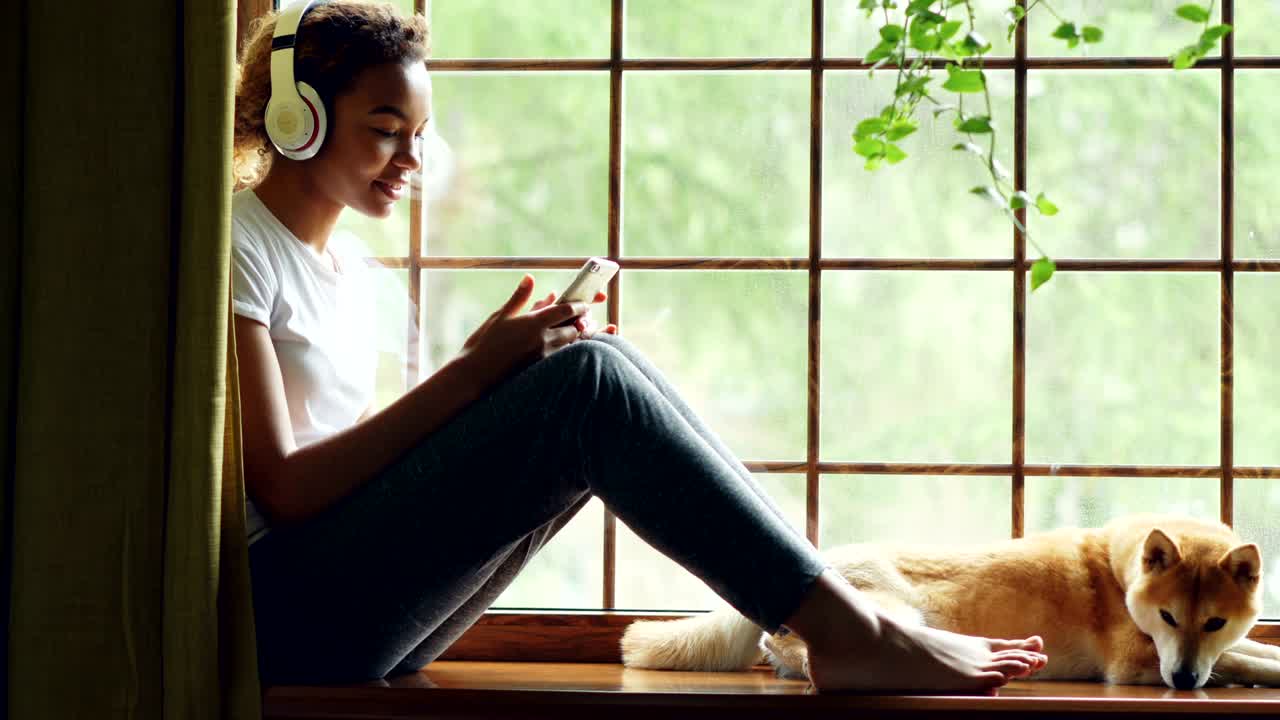 Pretty African American girl is listening to music with headphones and using smartphone sitting on window sill with lovely well-bred dog lying near her.