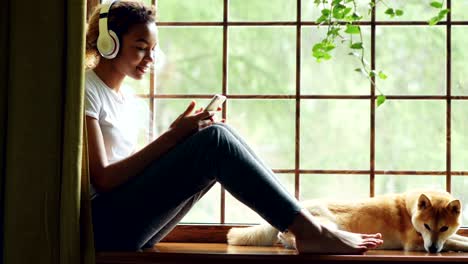 pretty african american girl is listening to music with headphones and using smartphone sitting on window sill with lovely well-bred dog lying near her.