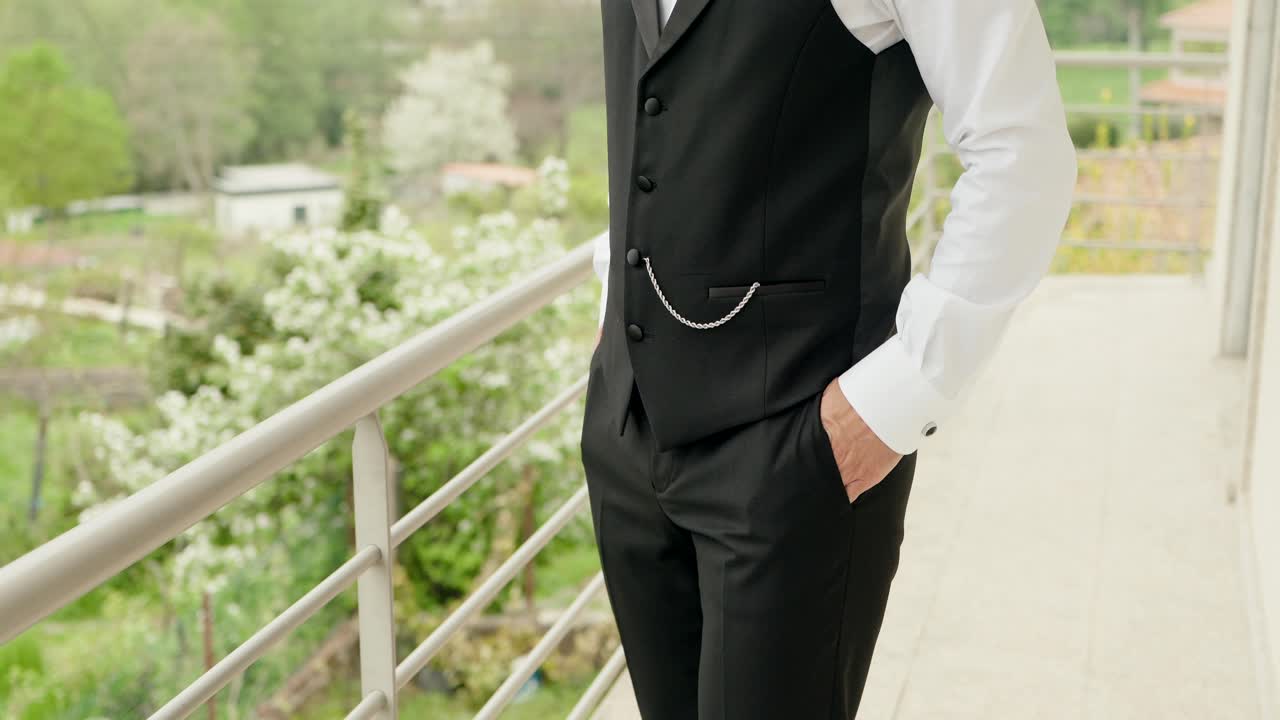 Close up of man in black vest and white shirt standing on balcony with hand in pocket