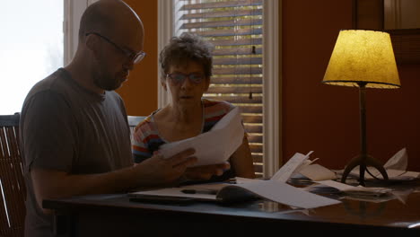 middle-aged man, and senior woman sit together at a dining table in a warmly lit home, sorting through papers. caregiver man helps older woman organize receipts, under table lamp light.